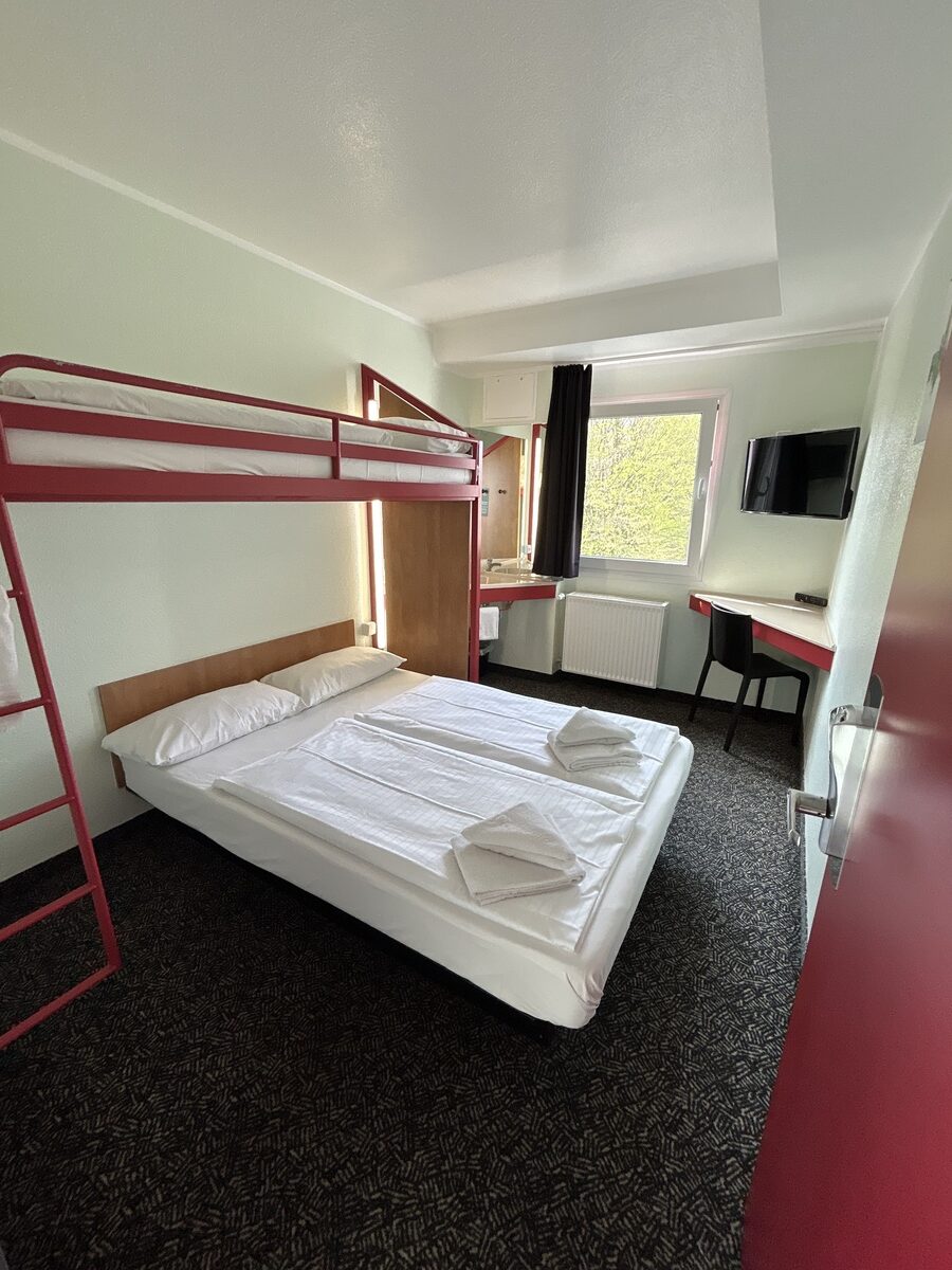 Zwei ungemachte Betten unter einem roten Hochbett in einem hellen Zimmer
