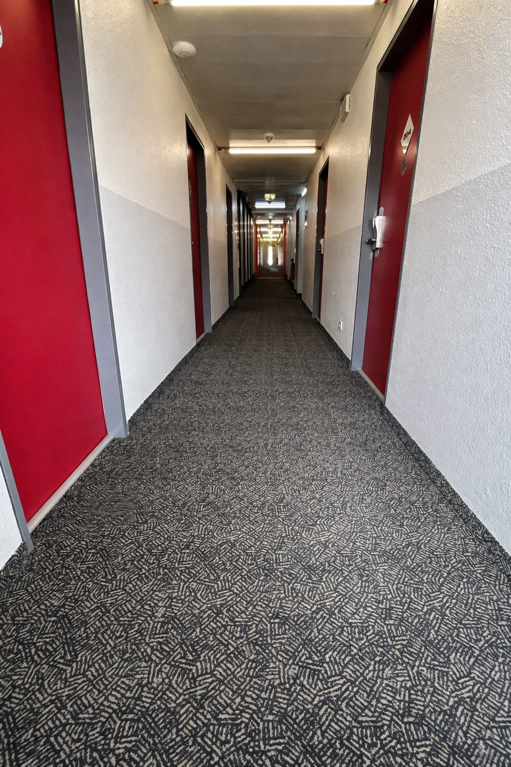 Langer Flur mit grauem Musterteppich und roten Türen, helle Beleuchtung