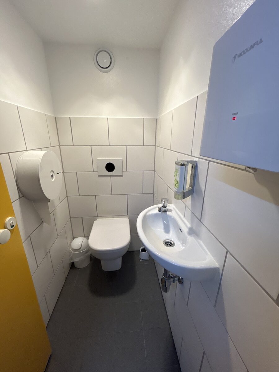 Eine kleine, weiß geflieste Toilettenkabine mit gelber Tür und Wandwaschbecken