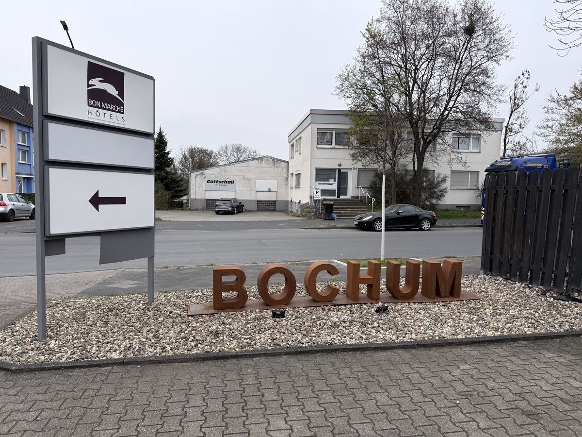 Ein großes, braunes BOCHUM Schild steht auf Kies vor einem Hotel und Bäumen