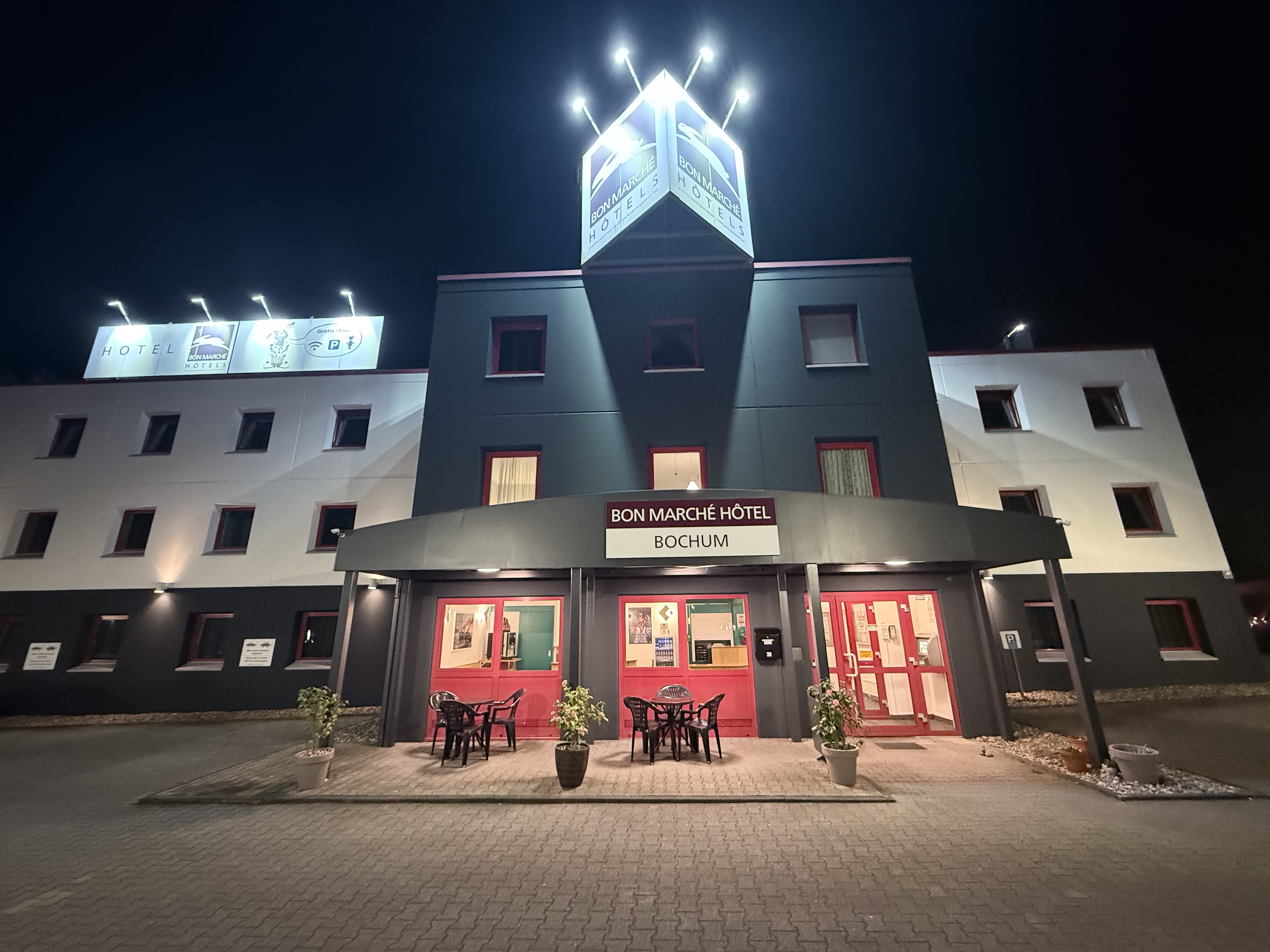Beleuchtetes Hotelgebaeude in Grau Weiss Rot mit markantem Turm bei Nacht